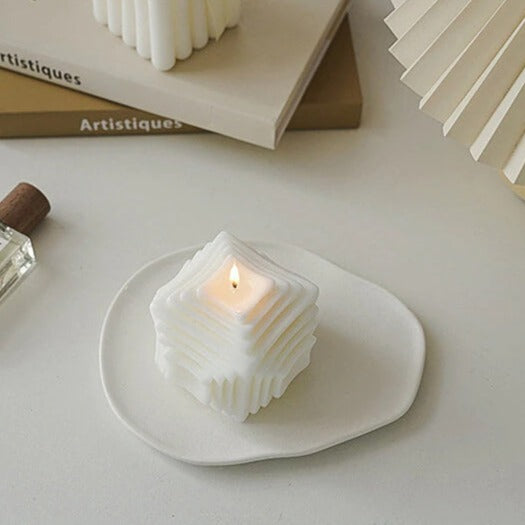 White, geometric candle on a white plate, with a lit flame.